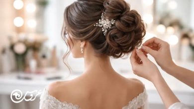 تصویر از پشتِ سرِ عروس در حال شینیون؛ آرایشگر در حال فیکس‌کردن مدل مو با سنجاق، شینیون جمع و حجیم با اکسسوری مرواریدی، لباس یقه باز و نور نرم آتلیه در پس‌زمینه.